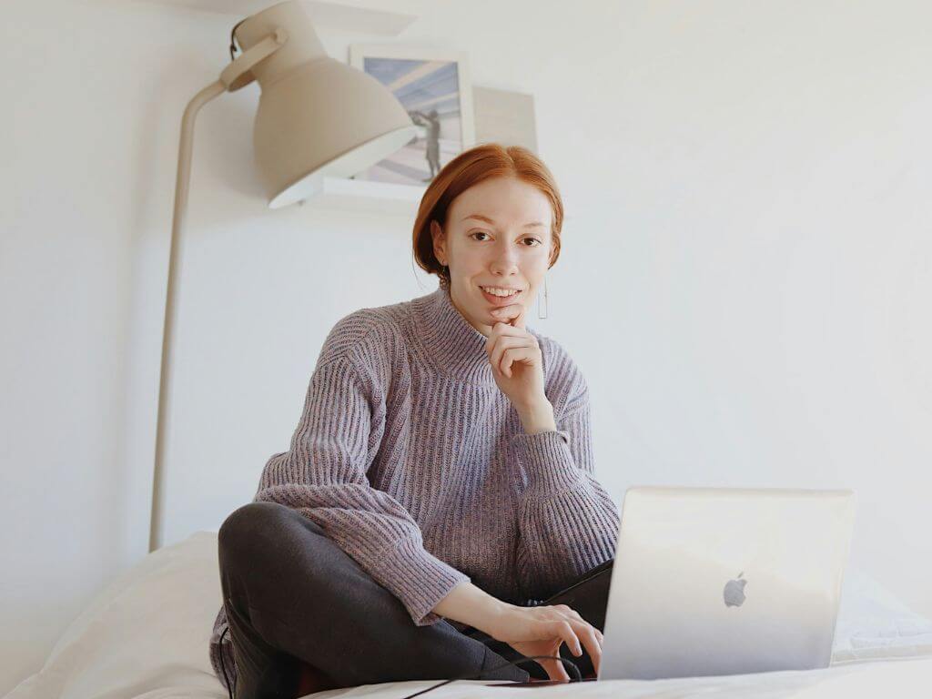 A young woman freelancer sitting on a bed with a laptop, smiling at the camera.