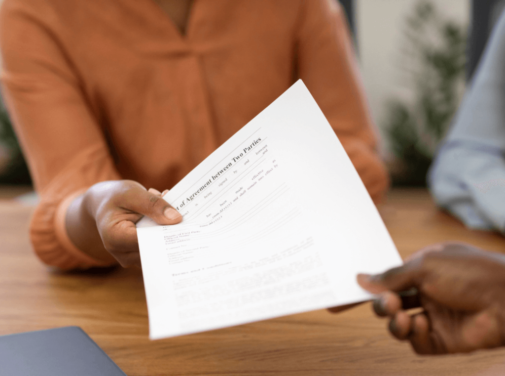 Close-up of a person handing over a printed agreement across a desk.