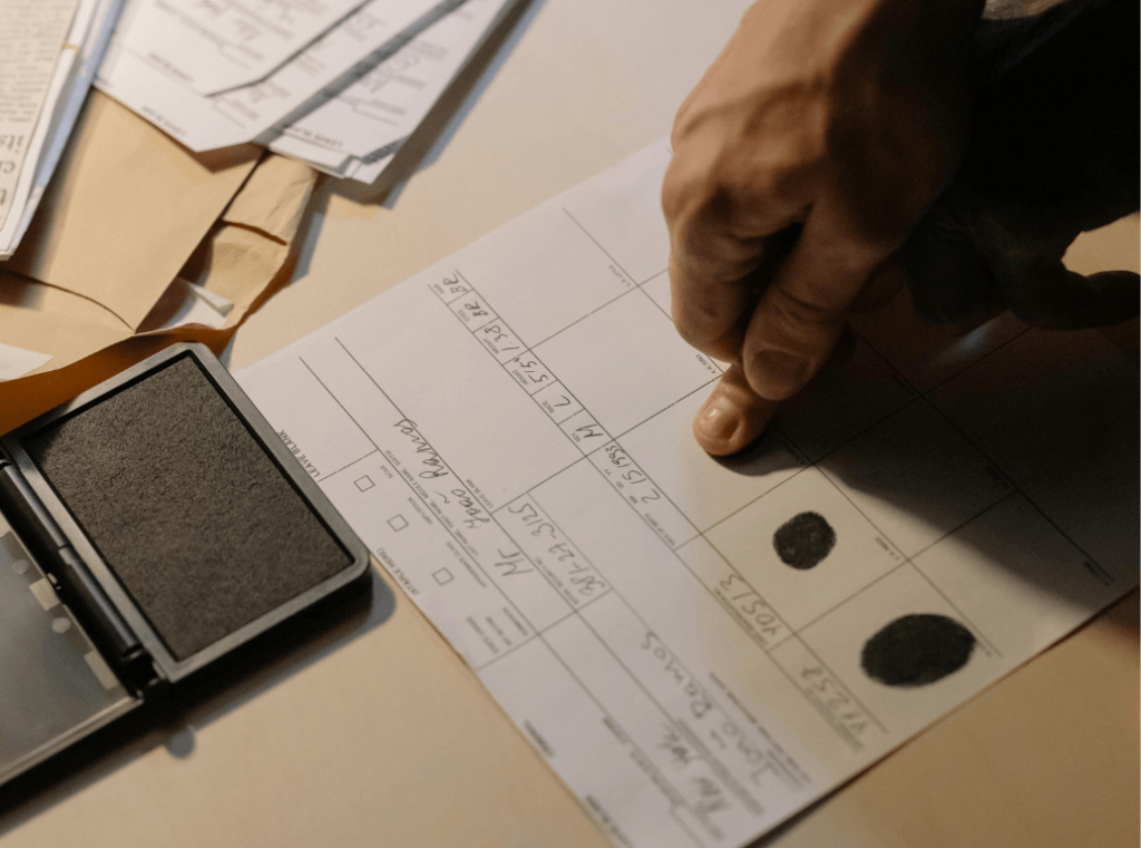 Hand pressing a finger onto a fingerprint card next to an ink pad, with documents in the background.