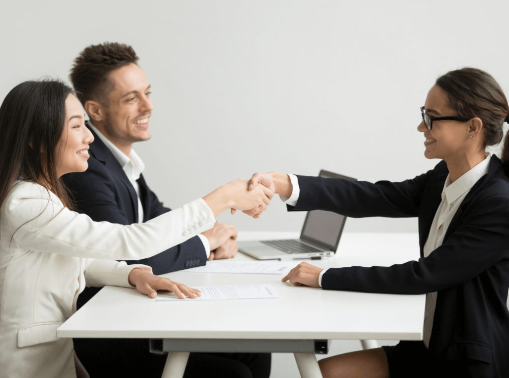 Smiling woman shaking hands with an interviewer across a desk, with another interviewer sitting beside them.