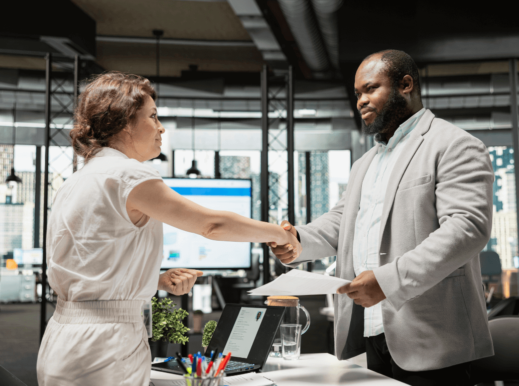 A professional woman and man shaking hands across a desk during an interview.