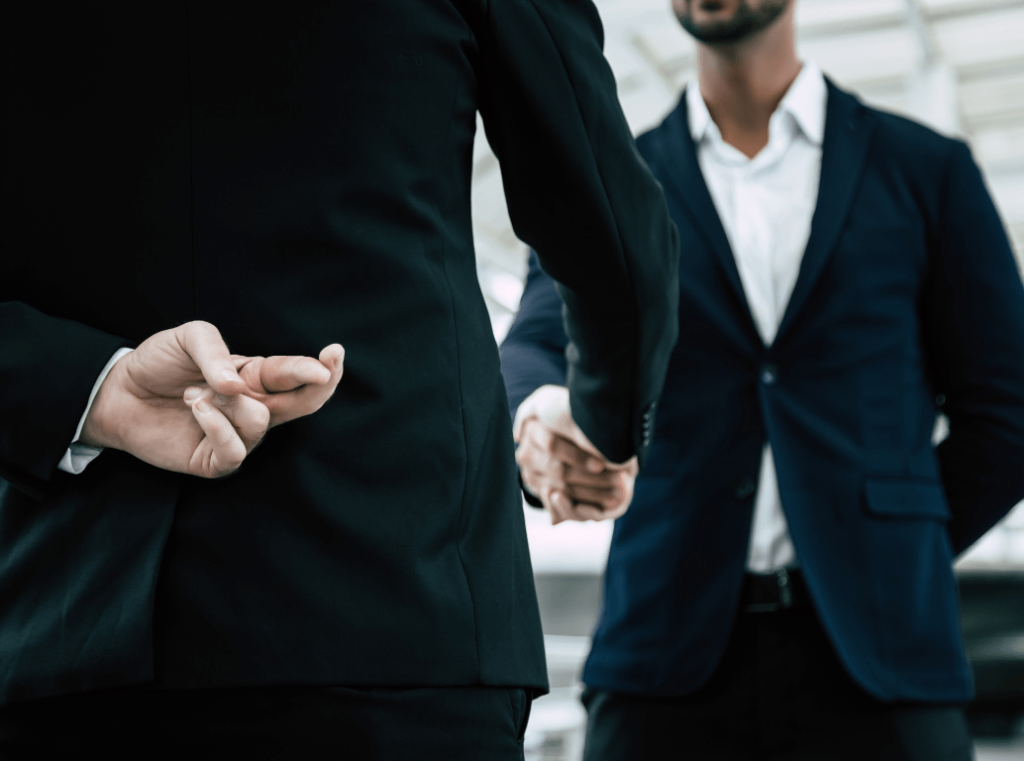 A close-up of a professional handshake between two individuals in suits, with the person in the foreground crossing their fingers behind their back to symbolise dishonesty.