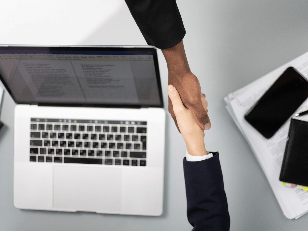A business handshake over a laptop and documents, representing a successful interview, contract signing, or hiring decision.
