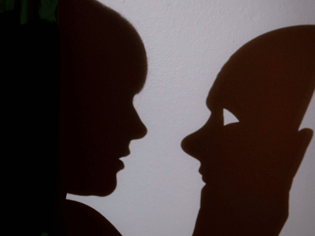 Silhouetted shadow of a person's profile facing a mask on a wall.